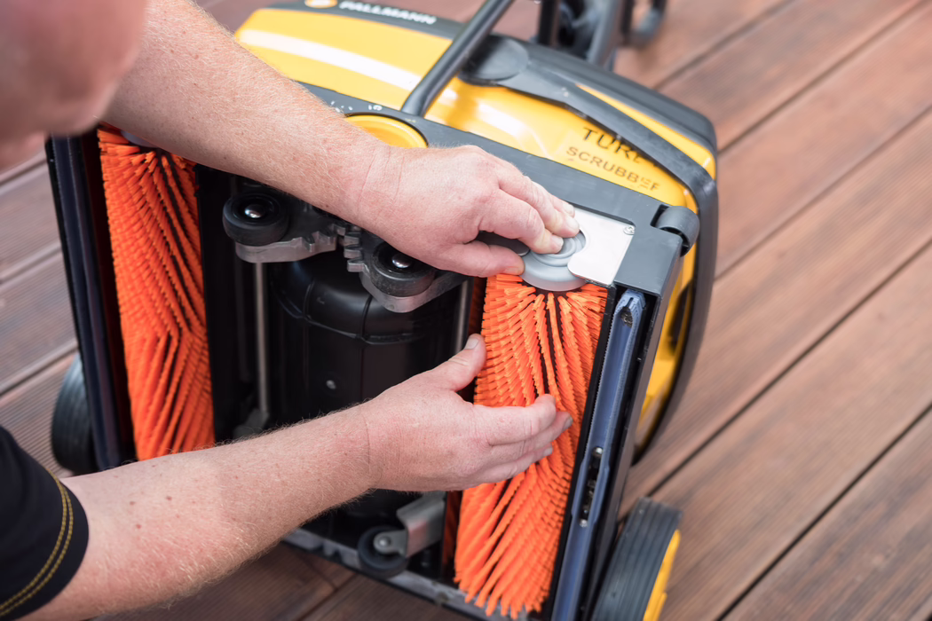 Nahaufnahme von Händen einer Person, die leuchtend orangefarbene Zylinderbürsten an der Unterseite einer gelben Bodenreinigungsmaschine auf einem Holzdeck anpasst.
