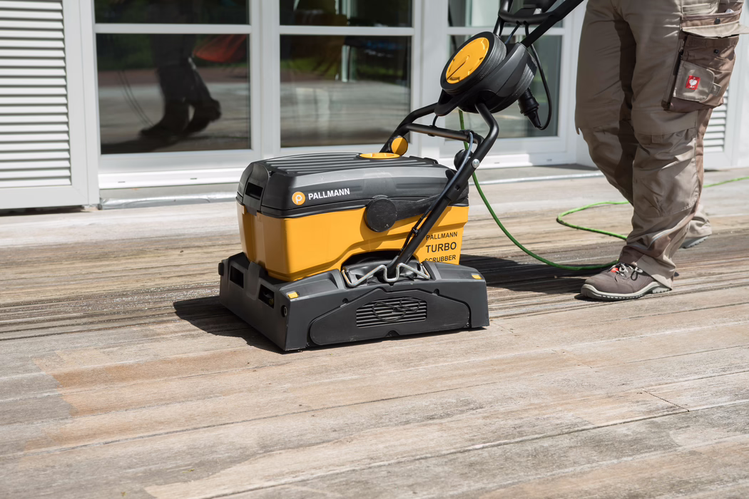 Ein gelb-schwarzer Pallmann Turbo Bodenreiniger reinigt ein verwittertes Holzdeck vor verglasten Terrassentüren. Rechts sind die Beine eines Arbeiters in braunen Arbeitshosen und grauen Schuhen zu sehen, ein grünes Kabel führt zur Maschine, die eine gereinigte Spur im Holz hinterlässt.