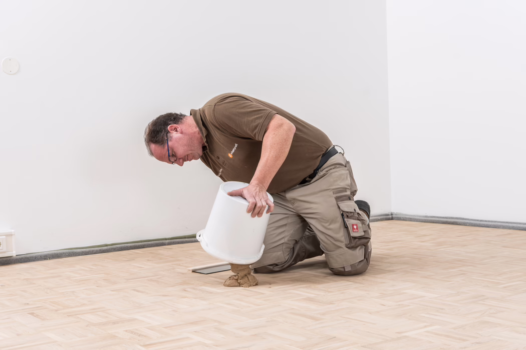 Ein Handwerker kniet auf einem frisch verlegten, hellen Holzparkettboden in einem weiß gestrichenen Raum und gießt sorgfältig eine beige Bodenmasse oder Klebstoff aus einem weißen Plastikbehälter auf den Boden, dabei hält er den Eimer mit beiden Händen; er trägt ein braunes Poloshirt, beige Arbeitshosen mit Knieschonern und Taschen sowie eine Brille.