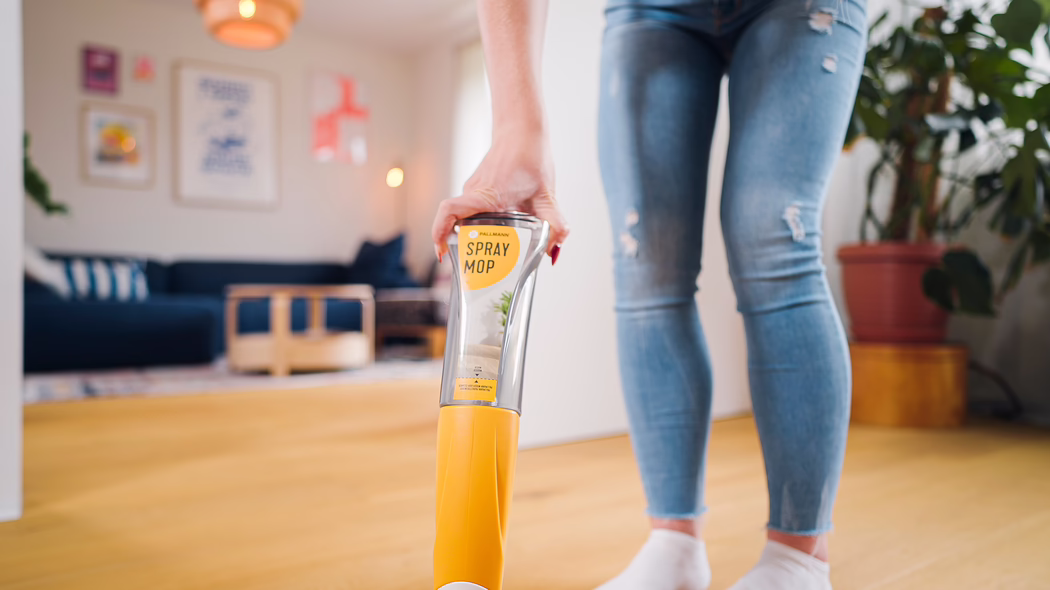 Pallmann_Application Image_Spraymop_072025 (24) Nahaufnahme einer Person, die mit einem gelben Spray-Mopp auf einem hellen Holzboden in einem modernen Wohnzimmer wischt. Zu sehen sind der Griff und der transparente Reinigungsflaschenbehälter mit der Aufschrift 'SPRAY MOP', gehalten von einer Hand mit rot lackierten Nägeln; die Beine der Person tragen zerrissene blaue Jeans und weiße Socken. Der weich verschwommene Hintergrund zeigt ein dunkelblaues Sofa, gerahmte Wandbilder, einen Holzcouchtisch, eine Topfpflanze und warme Lichtakzente, wodurch eine helle, saubere und gemütliche Wohnatmosphäre entsteht.
